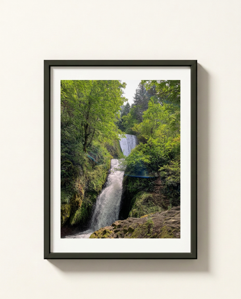 A framed landscape photograph of a lush green forest with a waterfall, presented in a black frame with a white mat against a light beige wall.