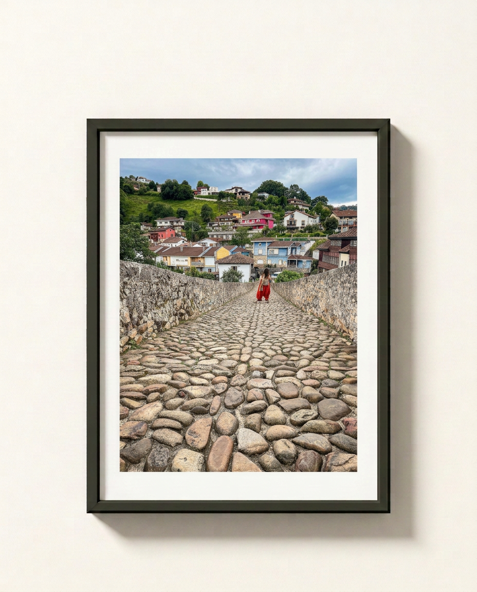 A framed photographic print of a cobblestone path leading up a hill toward a colorful village under a blue sky, with a person in red walking in the distance. The print is mounted with a white mat and enclosed in a black frame, hanging on a light beige wall.