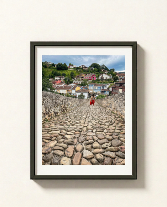 A framed photographic print of a cobblestone path leading up a hill toward a colorful village under a blue sky, with a person in red walking in the distance. The print is mounted with a white mat and enclosed in a black frame, hanging on a light beige wall.