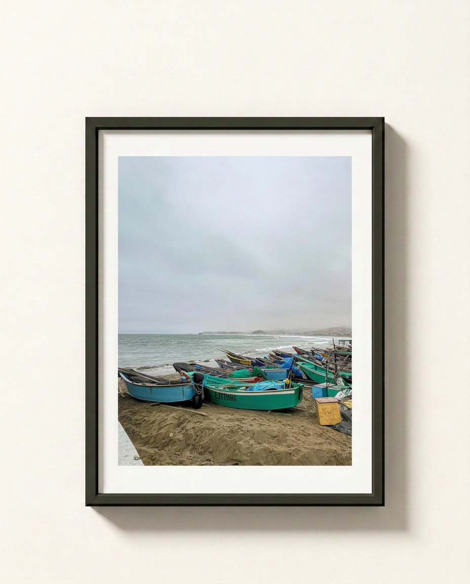 A framed photographic print of small colorful fishing boats resting on a sandy beach under an overcast sky, displayed on a light beige wall.