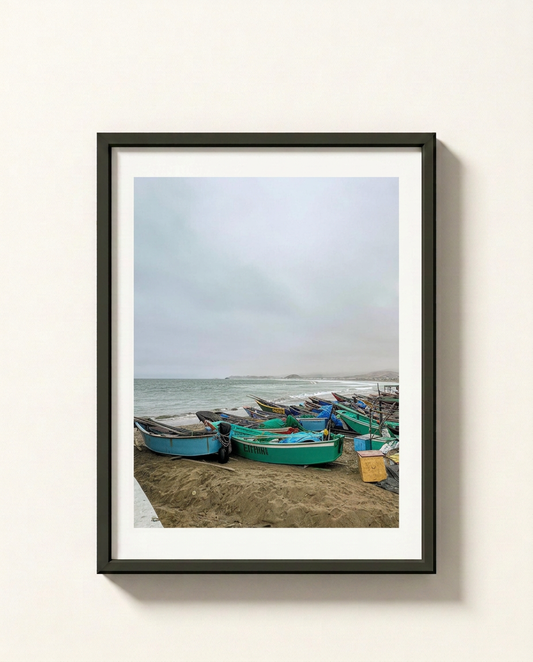 A framed photographic print of small colorful fishing boats resting on a sandy beach under an overcast sky, displayed on a light beige wall.