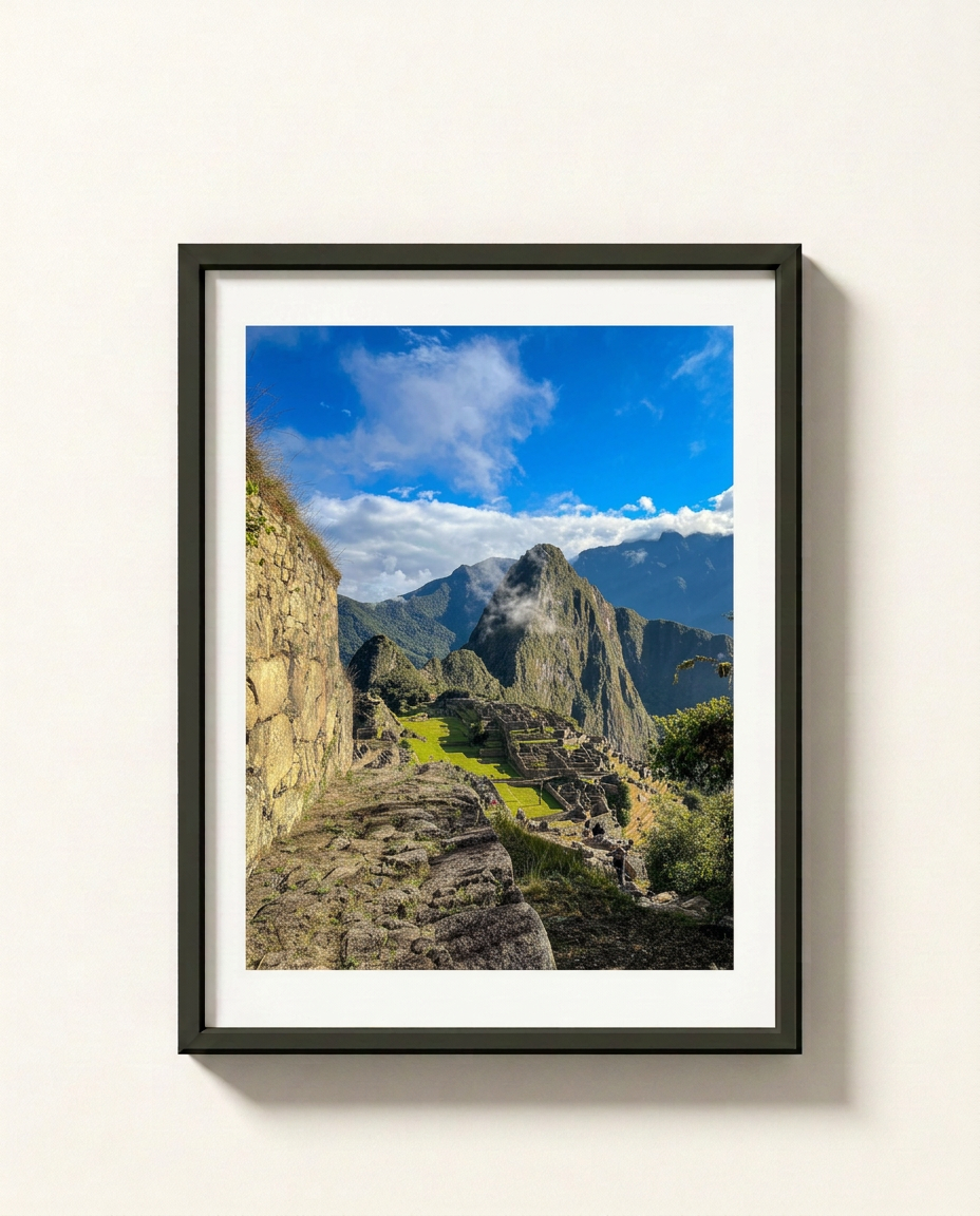 A framed photographic print of Machu Picchu, featuring the ancient stone ruins under a bright blue sky with white clouds, displayed on a clean off-white wall.