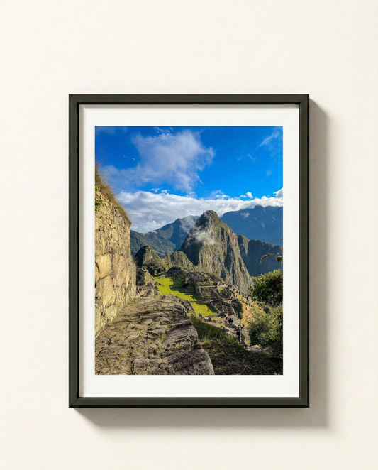 A framed photographic print of Machu Picchu, featuring the ancient stone ruins under a bright blue sky with white clouds, displayed on a clean off-white wall.