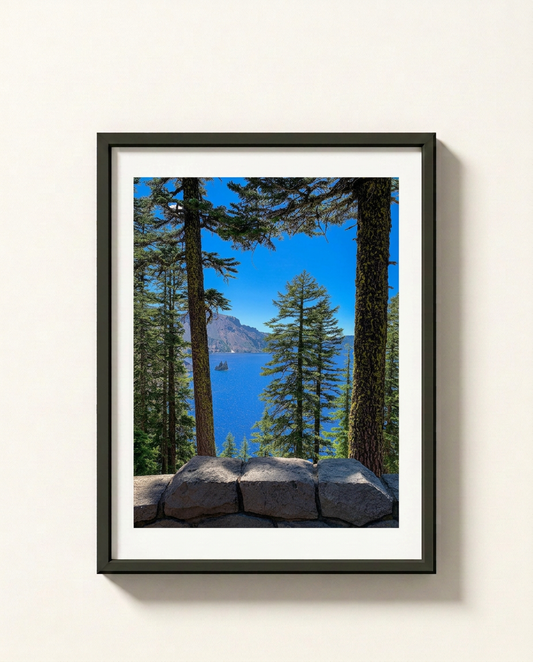 A framed landscape photograph of Crater Lake, Oregon, featuring a vibrant blue lake, tall pine trees, and a rocky foreground, presented in a black frame with a white mat against a light beige wall.