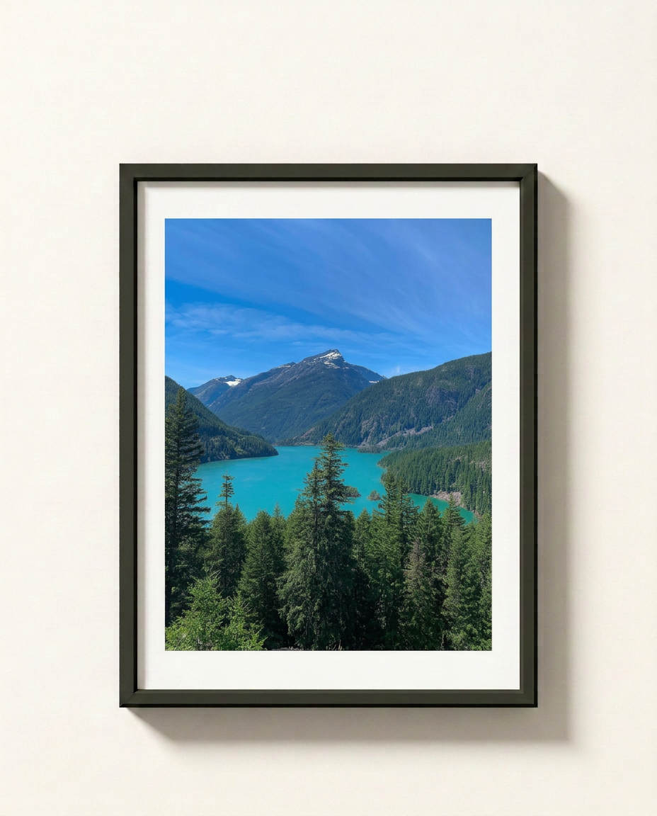 A framed landscape photograph of a turquoise lake surrounded by green mountains and pine trees under a blue sky, presented in a black frame with a white mat against a light beige wall.
