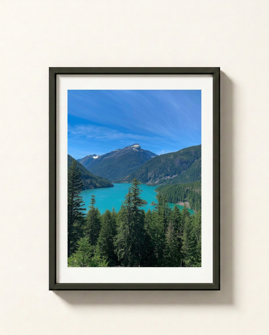 A framed landscape photograph of a turquoise lake surrounded by green mountains and pine trees under a blue sky, presented in a black frame with a white mat against a light beige wall.
