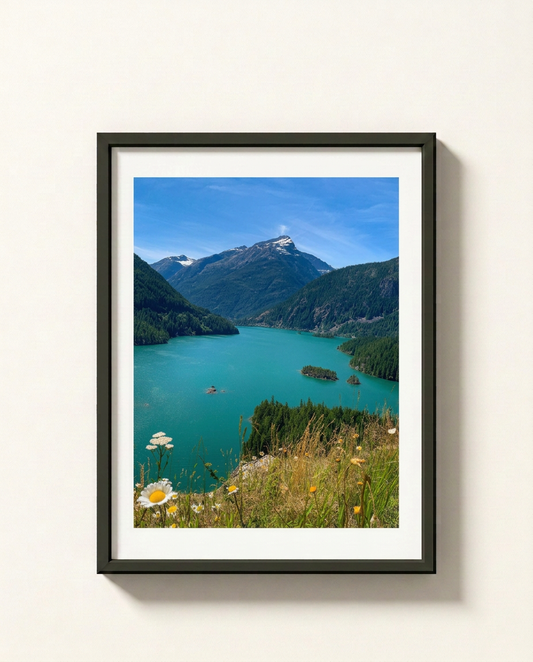 A framed landscape art print depicting a vibrant turquoise alpine lake surrounded by green mountains, a blue sky, and wildflowers in the foreground, mounted on a light beige wall.