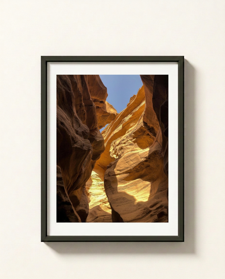 A framed vertical art print of a slot canyon with warm golden sandstone walls and a patch of blue sky, displayed on a light beige wall.