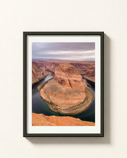 Framed landscape art print of Horseshoe Bend in Arizona, featuring the winding Colorado River around a red rock canyon under a cloudy sky, displayed on a light beige wall.