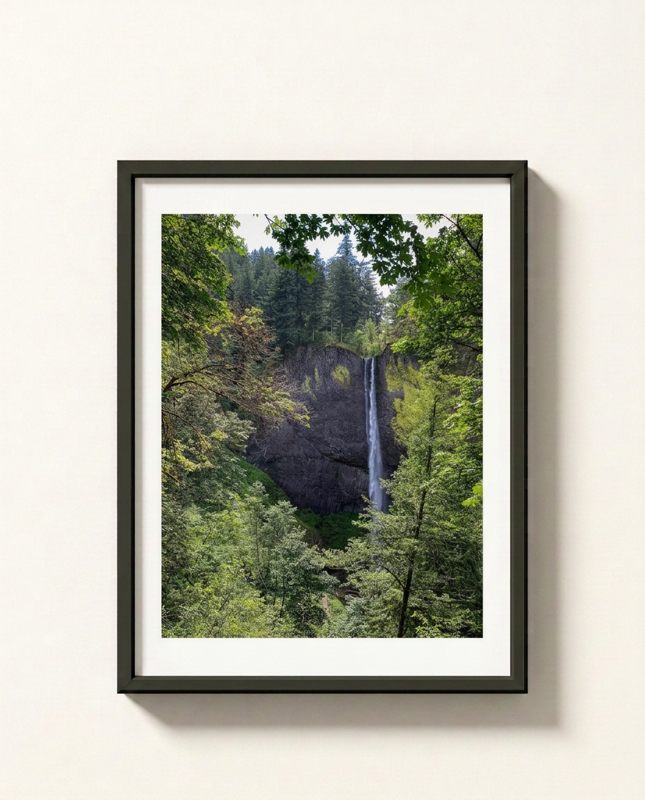 A framed landscape photograph of a waterfall cascading down a rocky cliff surrounded by lush green forest, presented in a black frame with a white mat against a light beige wall.