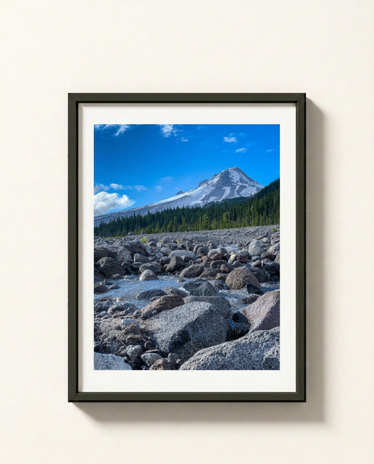 A framed landscape photograph of a snow-capped mountain under a bright blue sky, with a rocky riverbed and evergreen forest in the foreground, displayed on a light beige wall.