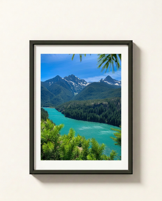 A framed landscape photograph of a turquoise river winding through a dense green forest and snow-capped mountains under a blue sky, presented in a black frame with a white mat against a light beige wall.