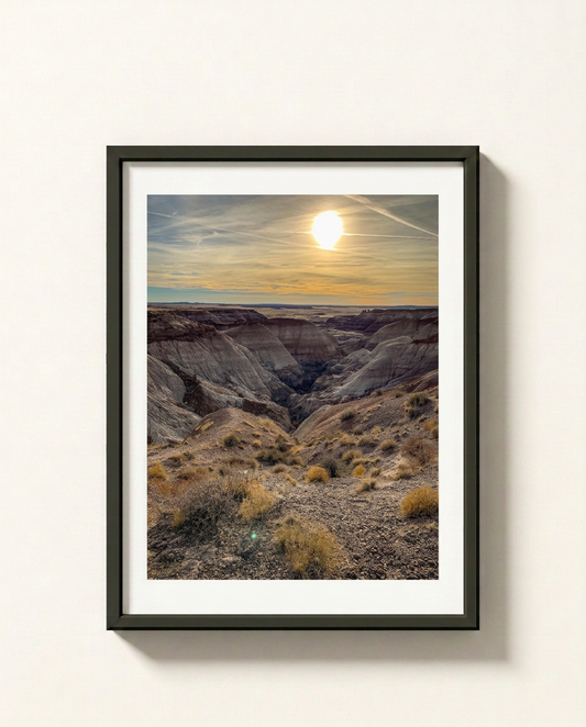 A framed landscape art print of a deep canyon under a glowing sun, displayed on a clean off-white wall. The print features a white mat and a sleek black frame.