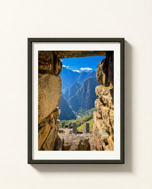 A framed art print of Machu Picchu, featuring a scenic view of the ancient ruins framed by stone walls under a blue sky, presented in a black frame with a white mat on a light beige wall.